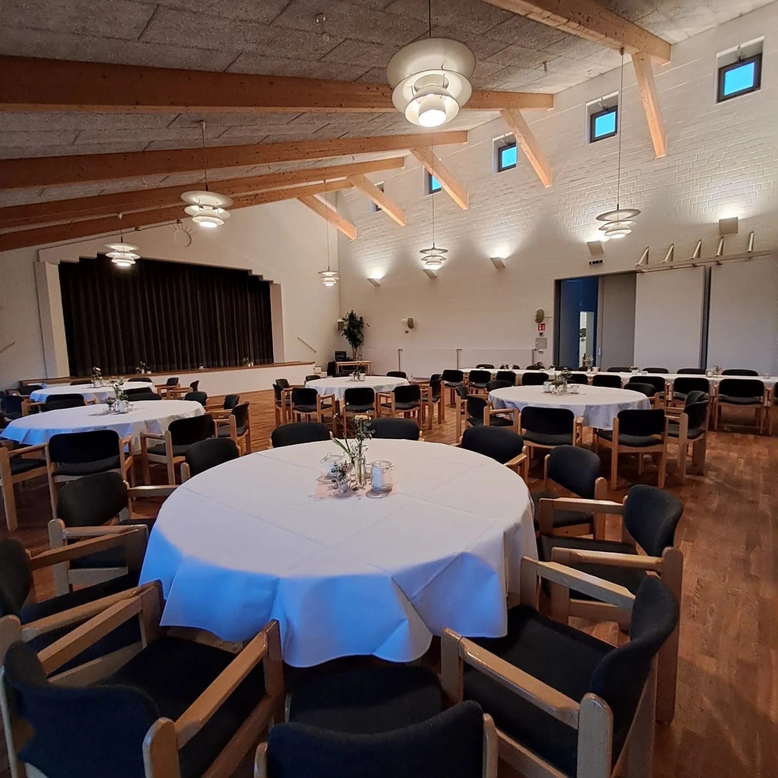 Derselbe Saal, für ein Fest gedeckt: runde Tafeln, Blumen auf jedem Tisch, Bühne im Hintergrund