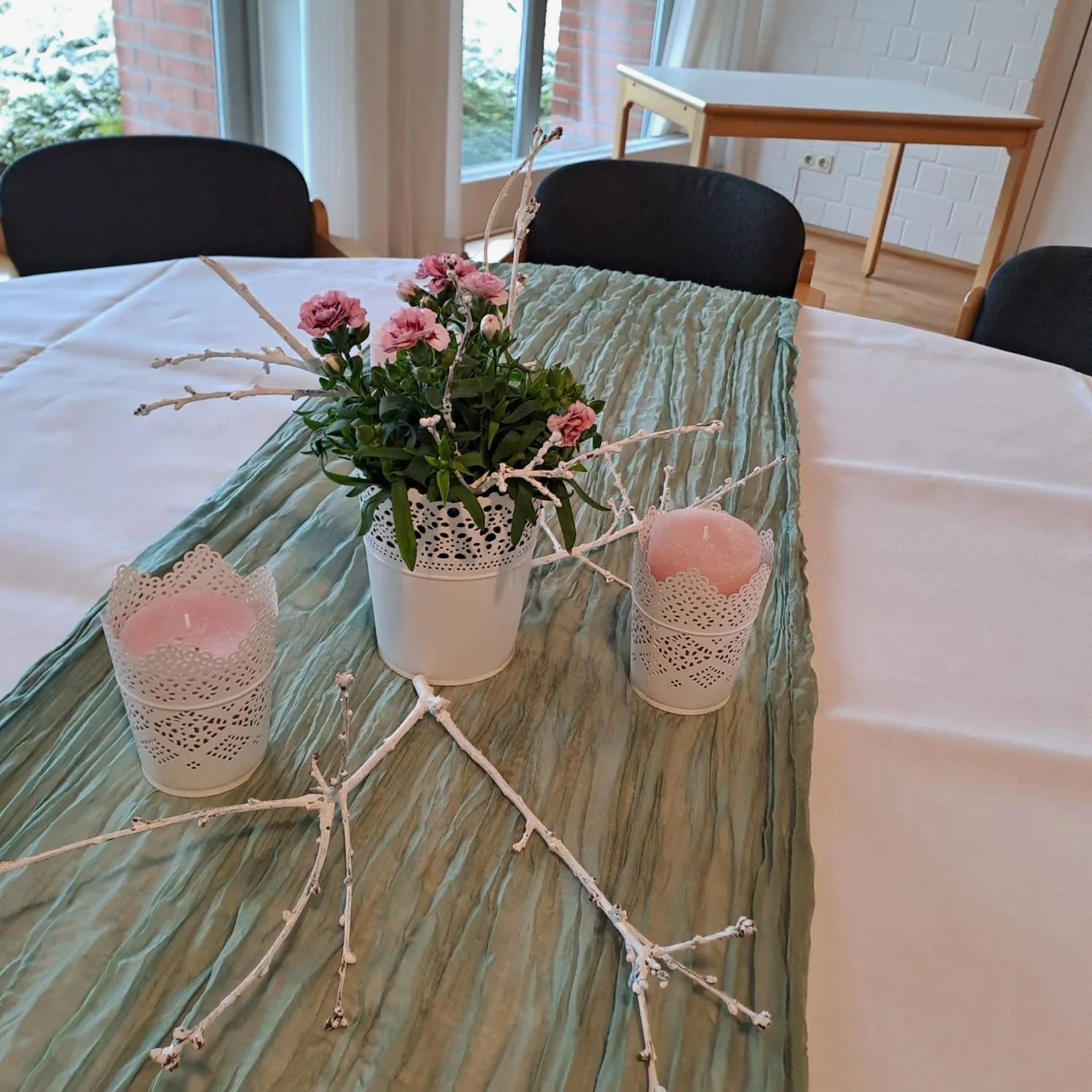 Tischschmuck aus rosa Rosen in weißen Übertöpfen, rosa Kerzen und einem weiß gekalkten Zweig auf einem salbeigrünen Läufer