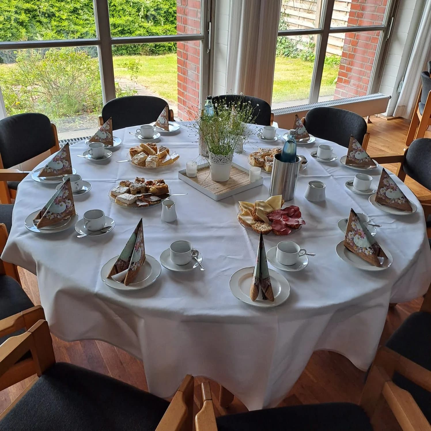 Runde Kaffeetafel am Fenster: Kuchenplatten, Tassen, gefaltete Servietten und Frühlingsgrün in der Mitte