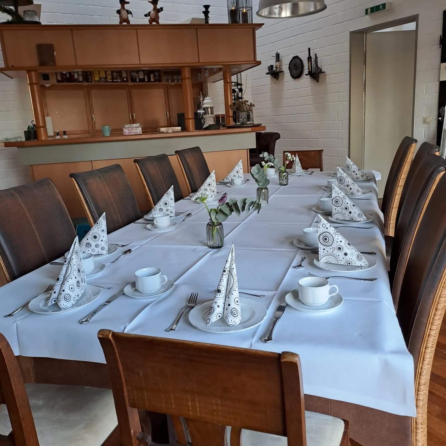 Schlicht gedeckte lange Tafel in der Gaststube: weiße Decke, Tassen, einfach gefaltete Servietten, dahinter der Tresen