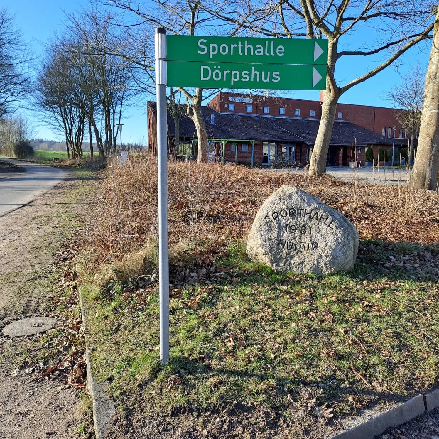 Grüner Wegweiser mit der Aufschrift Sporthalle und Dörpshus an der Straße, dahinter der Findling mit der Inschrift Sporthalle 1981 Hürup und das Haus