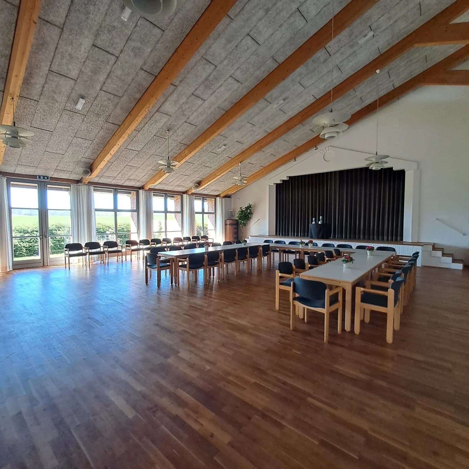 Der leere Saal bei Tageslicht: freies Parkett, Bühne mit dunklem Vorhang, Fensterfront zu den Feldern